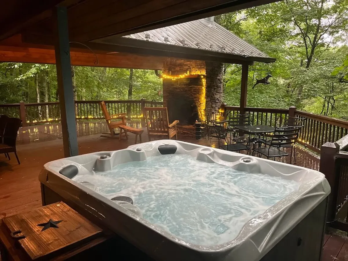 A tranquil wooden deck surrounded by lush greenery, featuring a bubbling hot tub and a cozy outdoor fireplace. The setup evokes a sense of relaxation and offers space for intimate gatherings or peaceful solo moments, with the hot tub as a central highlight.