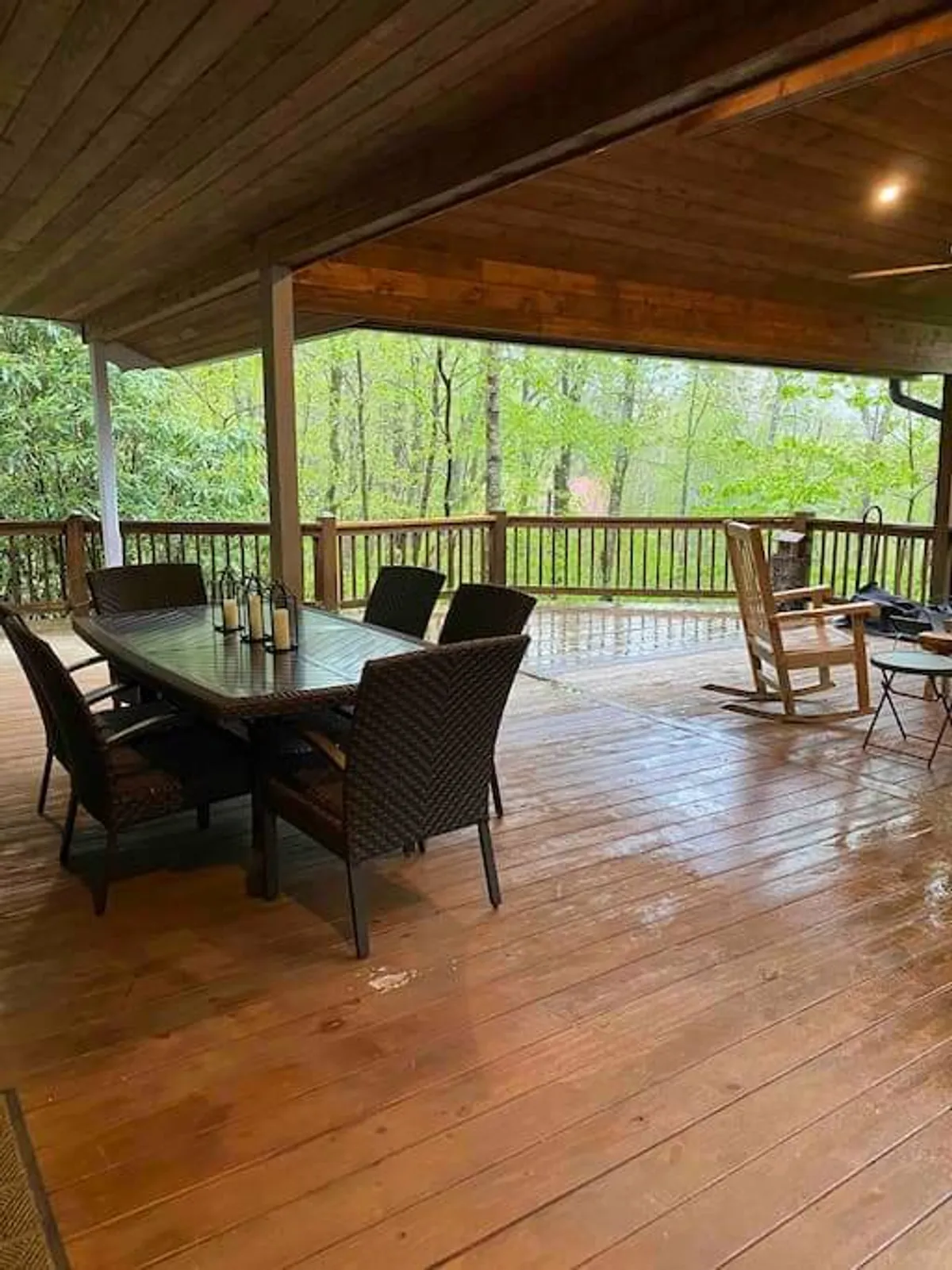 A wide wooden deck bordered by lush greenery, featuring a large outdoor dining table and a classic rocking chair. The setting is tranquil, inviting guests to unwind as evening falls or enjoy nature in the early morning. The deck’s spaciousness and natural surroundings create an ideal environment for both intimate and group gatherings.