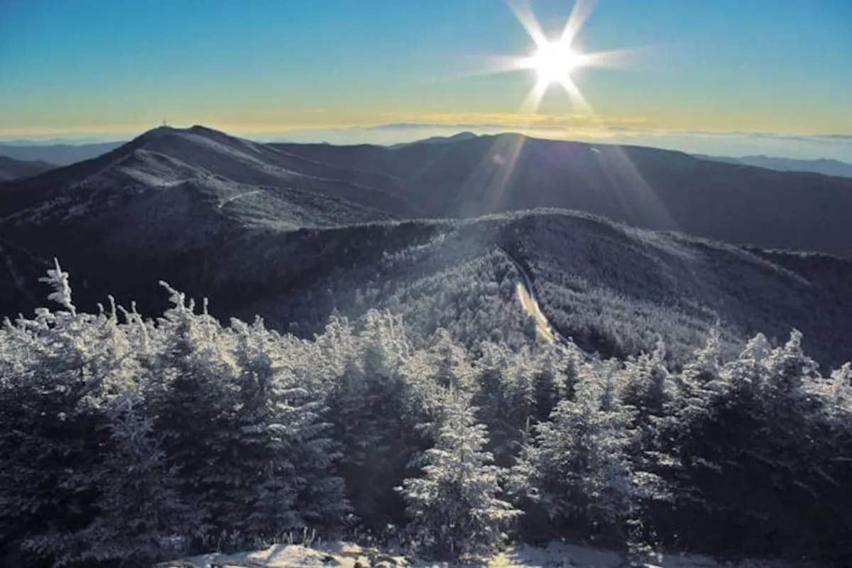 Snow-capped mountain peaks glow in early sunlight above dense evergreen forests, evoking the grandeur and seasonal beauty of the Appalachian mountains. The scene is tranquil, with a sense of boundless adventure and breathtaking vistas.