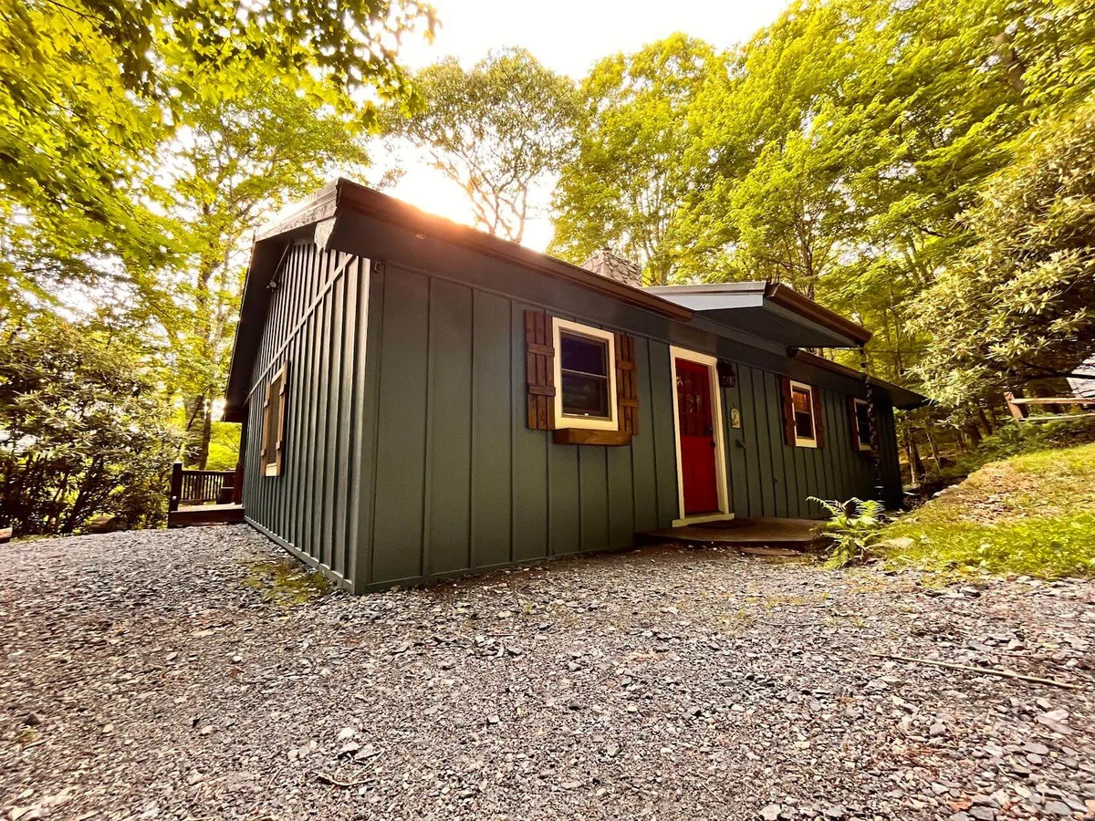 A welcoming mountain home with green exterior walls, a bright red door, large windows, and mature trees all around. The setting is cozy and exclusive, reminiscent of a private, gated resort community.