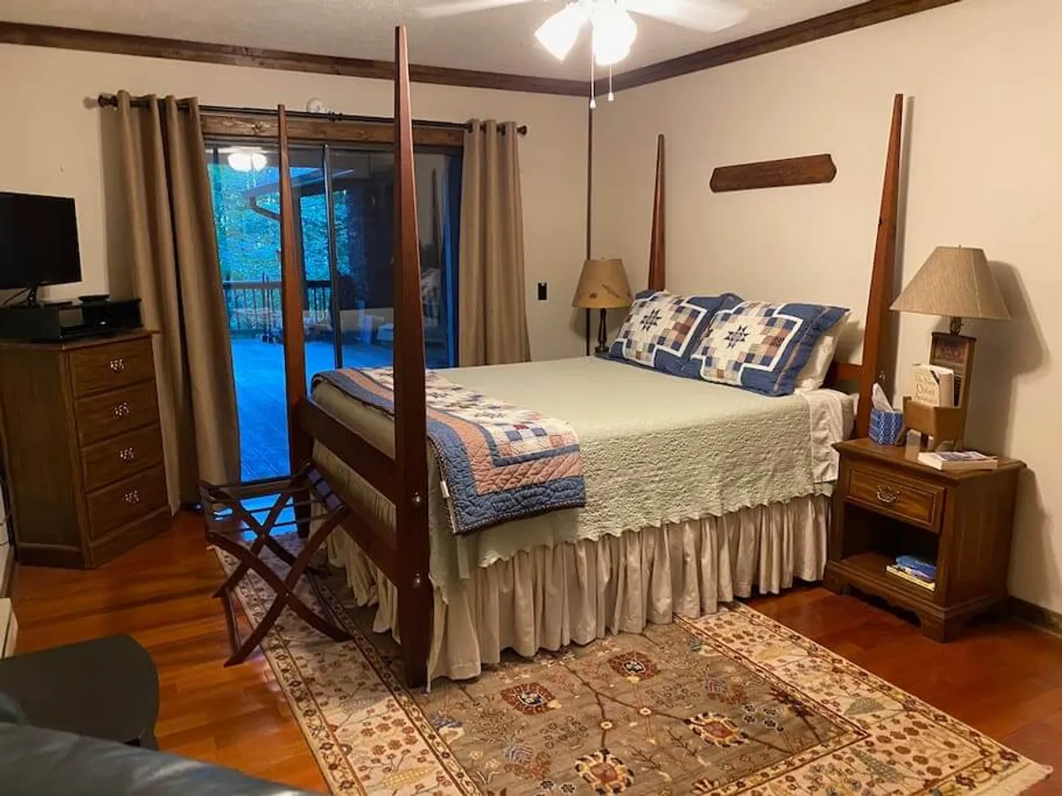 A cozy interior shot of a four-poster bed with a nightstand and lamp, an area rug, and a large window with curtains. The room feels warm and inviting, ideal for restful sleep and peaceful moments alone or with a loved one.