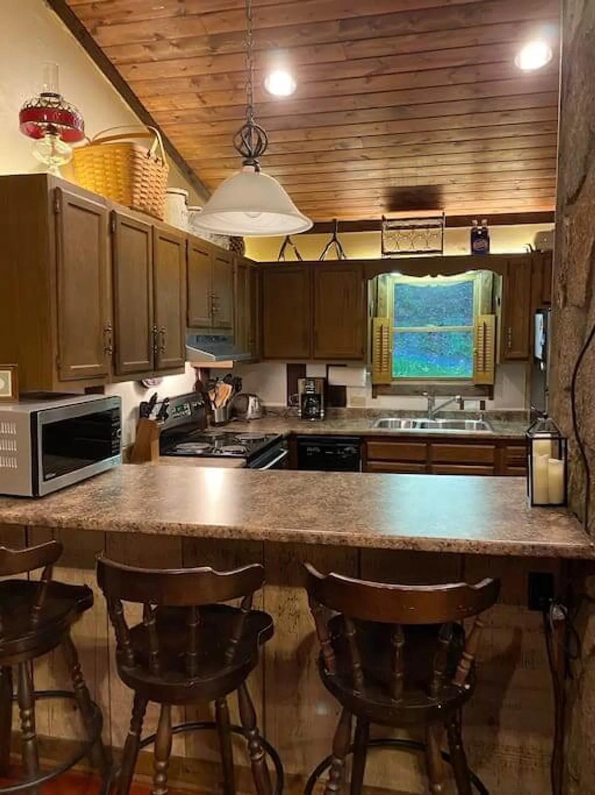 A bright kitchen area with wooden cabinets, a bar counter outfitted with stools, and a window offering an inspiring outdoor view. The bar counter invites casual gatherings and keeps the chef connected with the group while preparing meals.