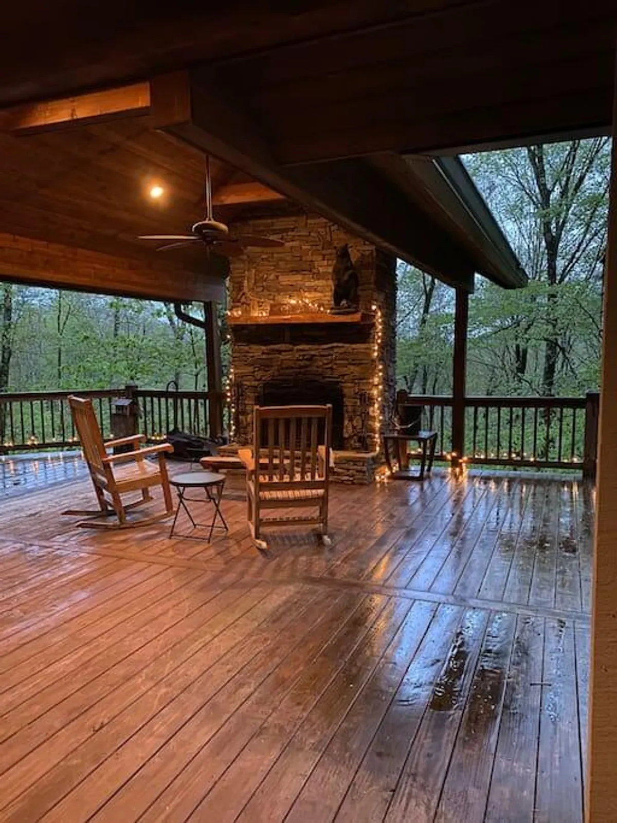 The image features a cozy mountain deck with a rustic stone fireplace, inviting wooden rocking chairs, and warm string lights twinkling overhead. The scene hints at comfortable relaxation, perfect for gathering with friends or sipping a hot drink while admiring the surrounding natural beauty.