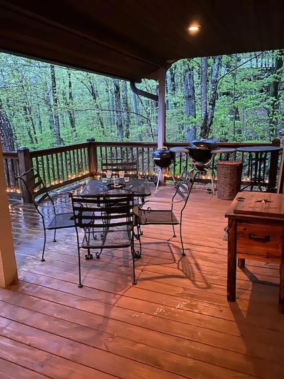 A wooden deck with a glass table and chairs, accompanied by a BBQ grill and bordered by a wooden railing. The scene is set for outdoor dining or casual gatherings, perfectly capturing the spirit of enjoying meals, drinks, and fresh mountain air on the deck.