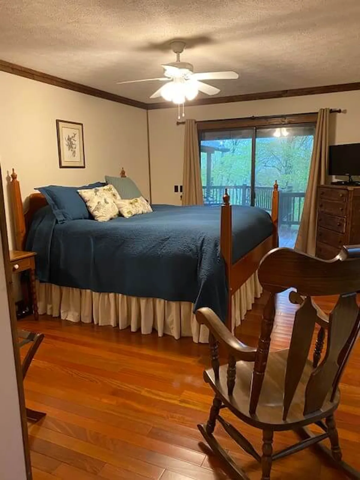 A tranquil bedroom featuring a bed with blue bedding, a rocking chair beside a large window with a view, and soft natural light. The space exudes calm and offers a cozy corner perfect for reading or quiet reflection.