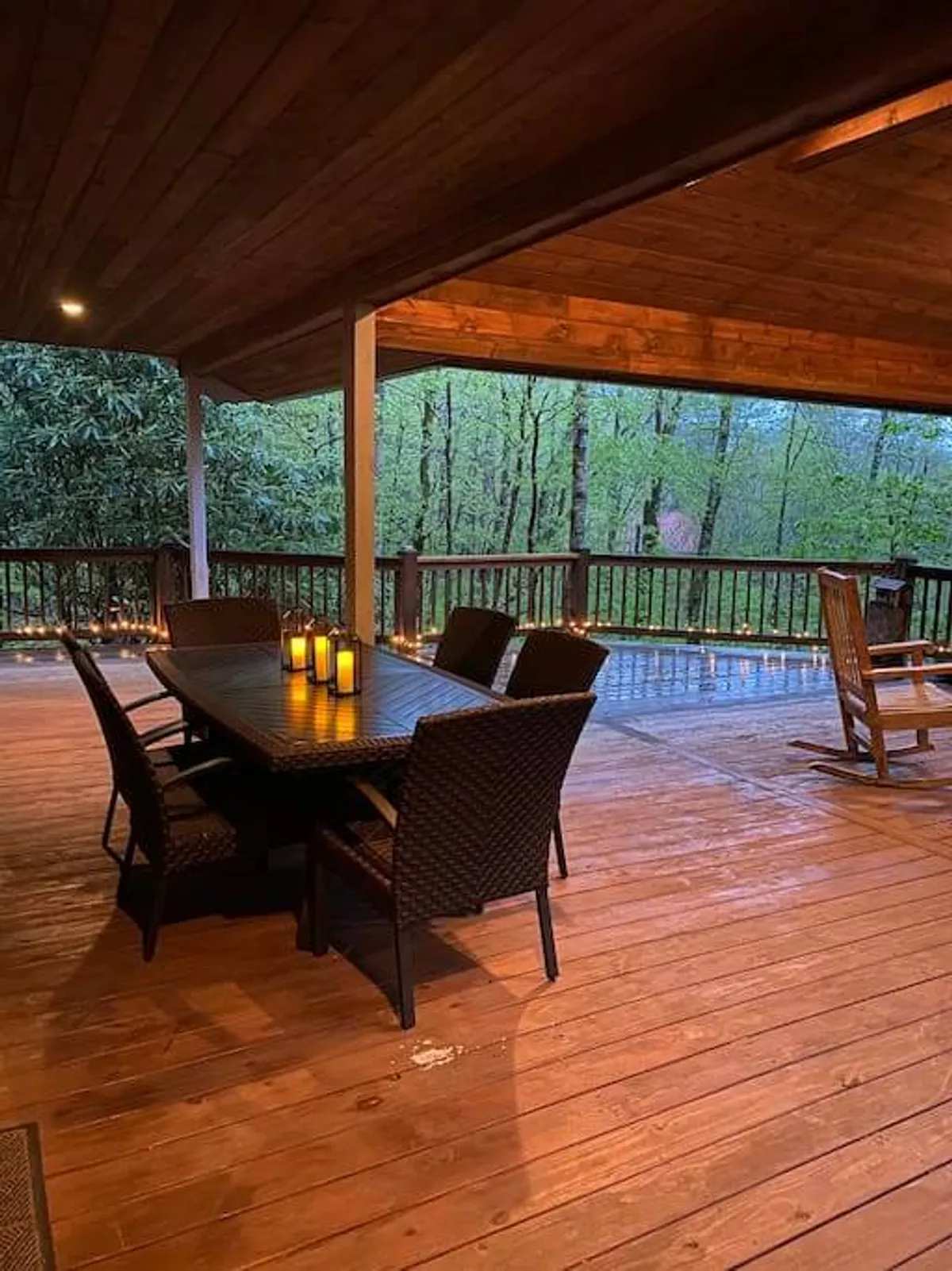 A serene exterior deck framed by tall trees, featuring a wooden dining table set with candles and a comfortable rocking chair. The setting is intimate, perfect for candlelit dinners, stargazing, or quiet reflection in nature’s embrace.